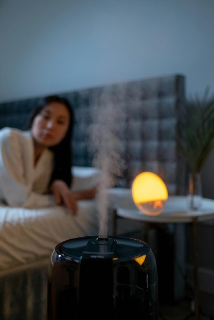 An Asian woman in pajamas rests in a bedroom with a humidifier and warm lighting, creating a cozy ambiance.